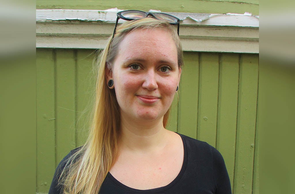 Porträtt av Nina Nyman, blond med långt hår, glasögon på hjässan och svart topp framför en grön brädvägg.